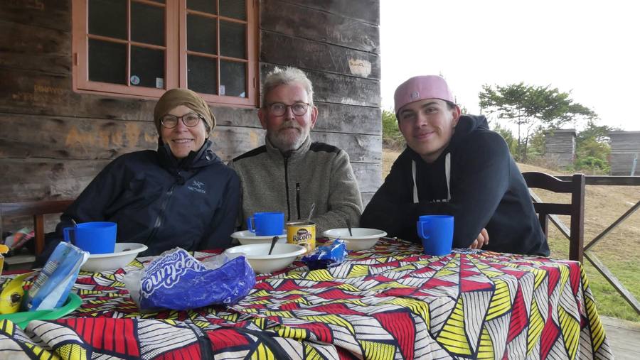 Three visitors sit at a patterned tablecloth outside a timber mountain hut with mugs and bowls of breakfast.