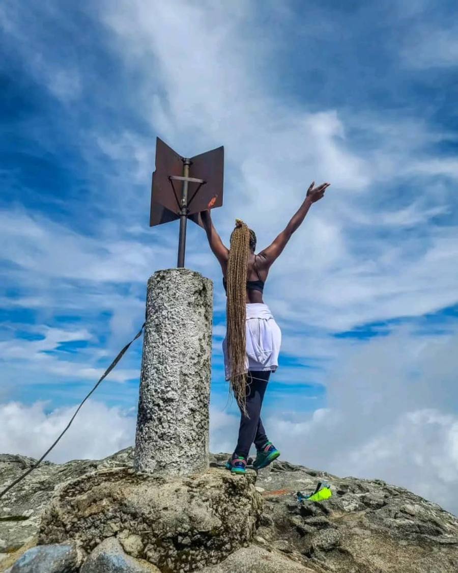 A walker standing at the summit marker on Chilemba peak with one arm raised, against a blue sky and cloud.
