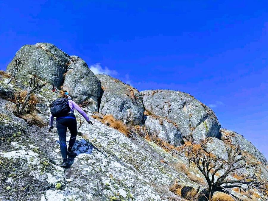 A walker with a backpack scrambling up a lichen-covered granite slab on the way to Sapitwa Peak.