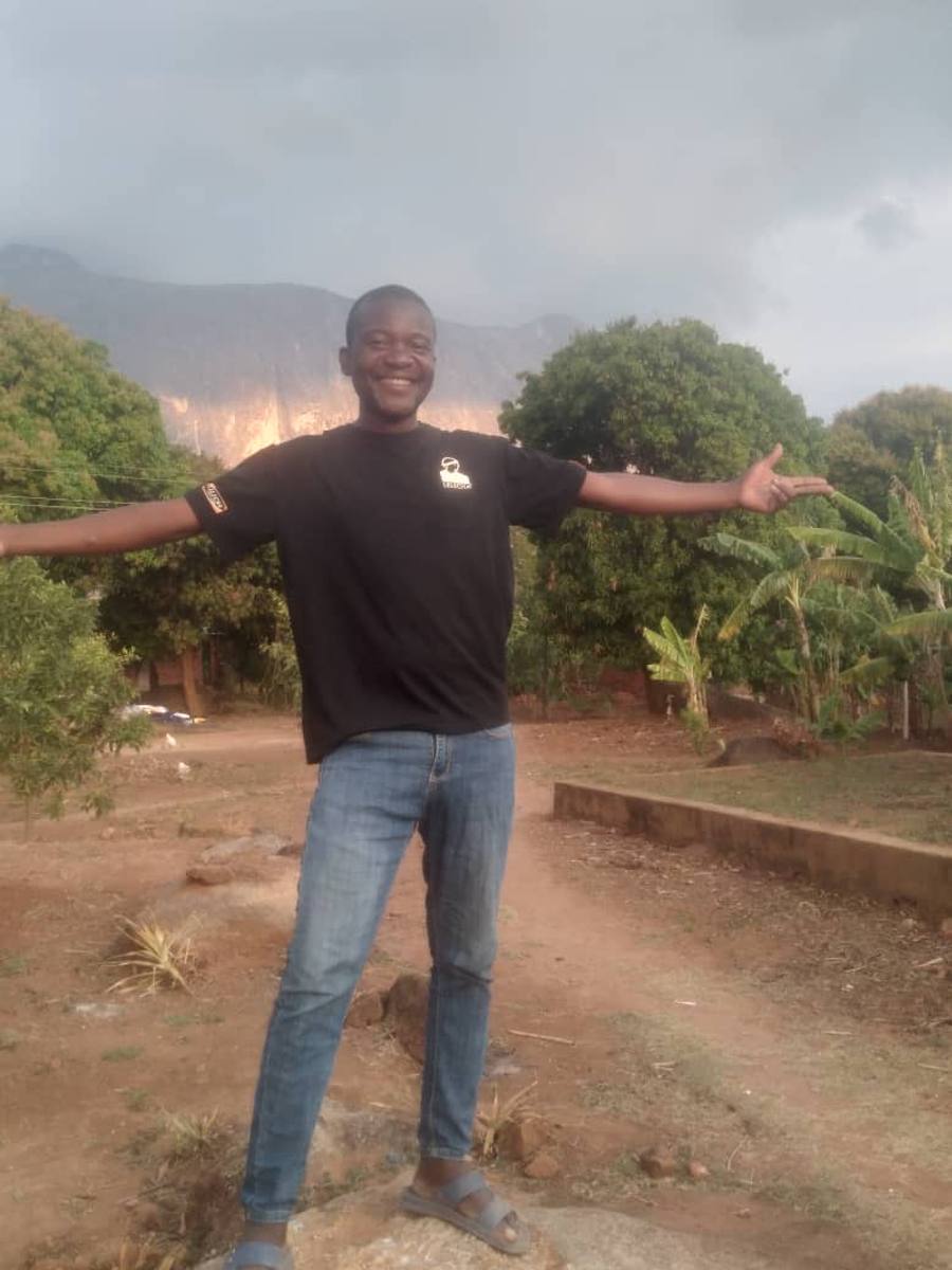 Edgison Chimowa smiling, crouched on a boulder in a garden of mango and banana trees, with the Mulanje massif rising behind him.