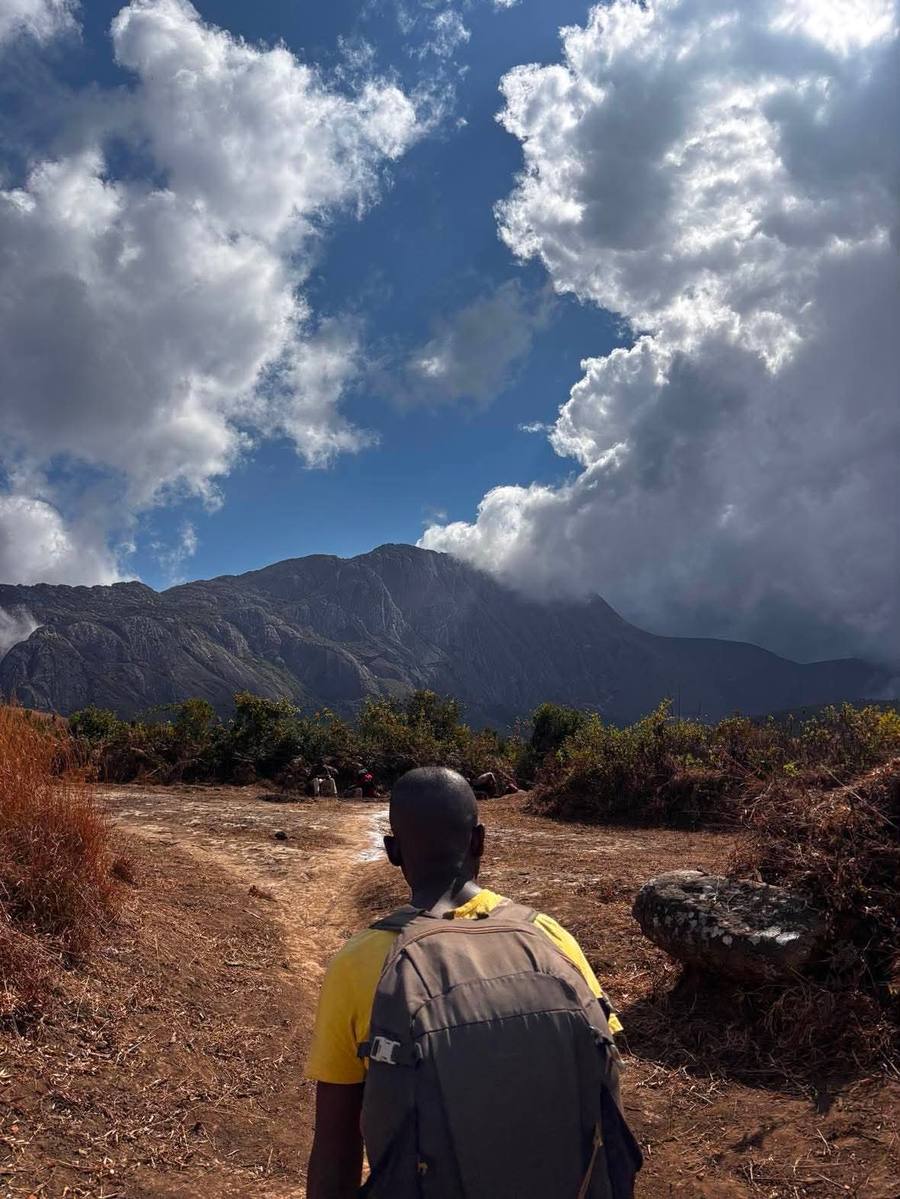 A guide with a backpack walks a dirt path towards the Mulanje massif under tall cumulus cloud.