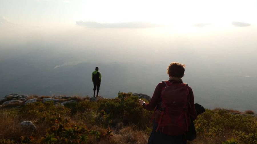 A hiker stands at the edge of the plateau looking out over haze, with a second walker in the foreground.