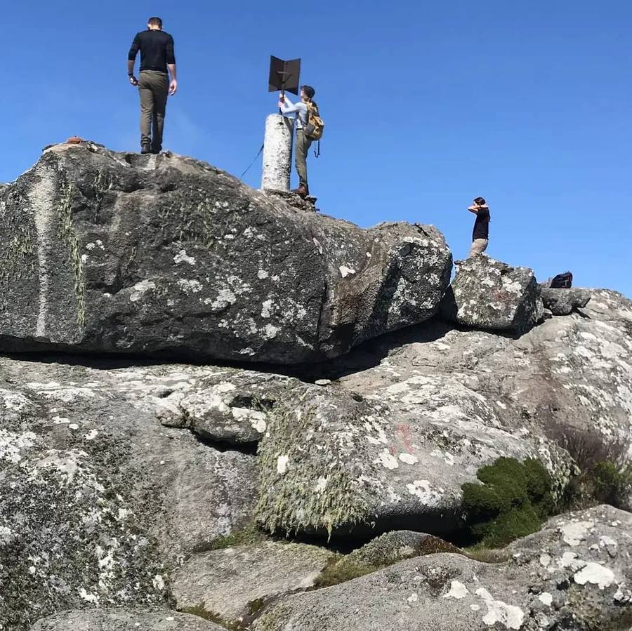 Walkers on the boulders at the summit of Sapitwa Peak beside the summit marker, under a clear blue sky.