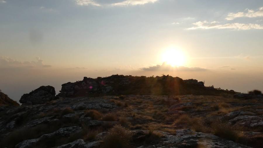 The sun rises over a rocky ridge on the plateau, backlighting tussock grass.