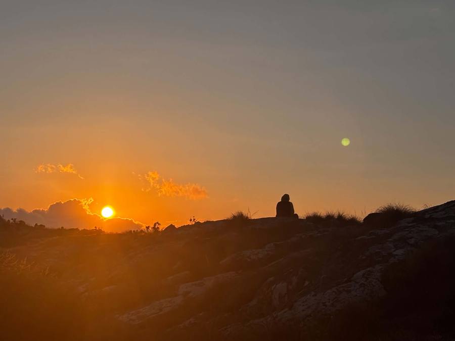A lone figure sits on a rocky plateau watching the sun set over the plain far below.