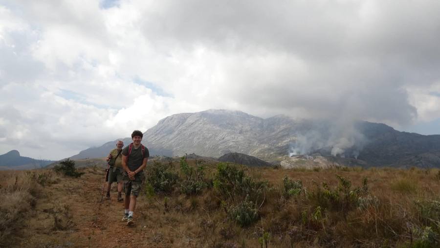 Two walkers on a grassy plateau path with the pale granite massif and drifting smoke behind.