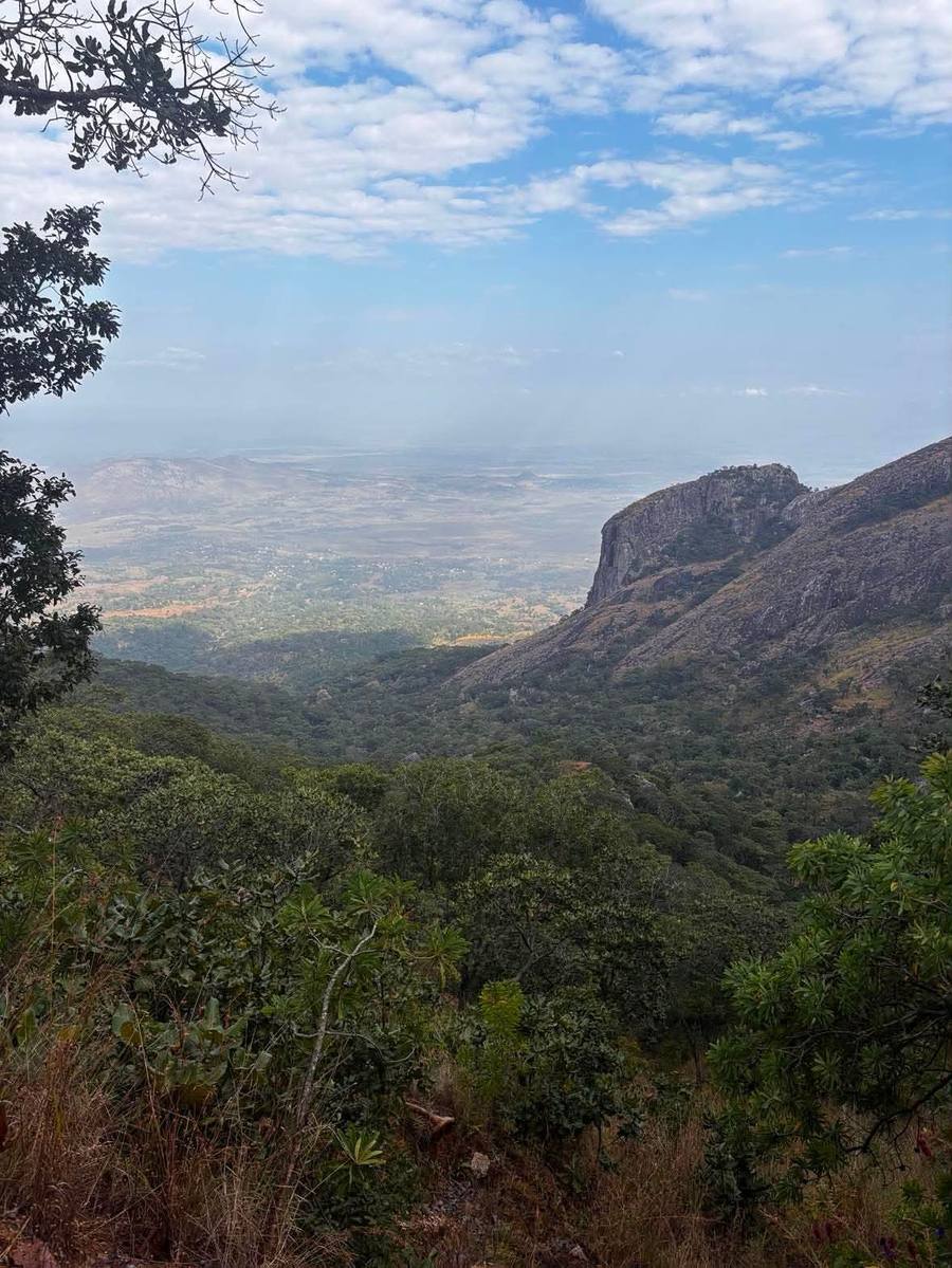 The plain far below seen from the mountain, with a rock buttress on the right and miombo woodland in front.