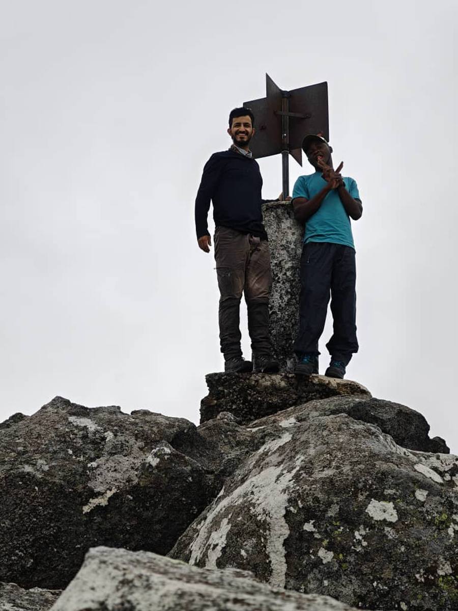 Two walkers standing beside a concrete summit marker on wet granite boulders in flat cloud.